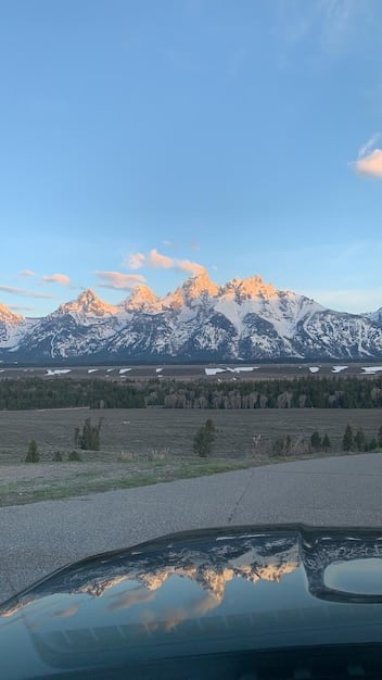 A modern SUV is parked at a scenic overlook, with a vast mountain range in the background under a clear blue sky. A person is checking the car's engine, with a focus on fluid levels and overall engine condition, conveying a sense of preparedness and vehicle maintenance on a road trip.