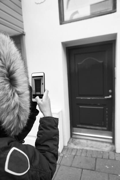 A close-up of a smart doorbell with a camera, displaying a live feed of a person approaching the door on a smartphone screen. The doorbell is mounted on a modern front door with a sleek design.