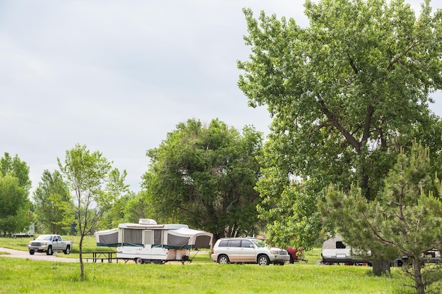 A well-maintained RV park with several RVs parked in designated spaces, surrounded by trees and greenery. The RVs are connected to utility hookups, and there are picnic tables and chairs set up outside each RV.