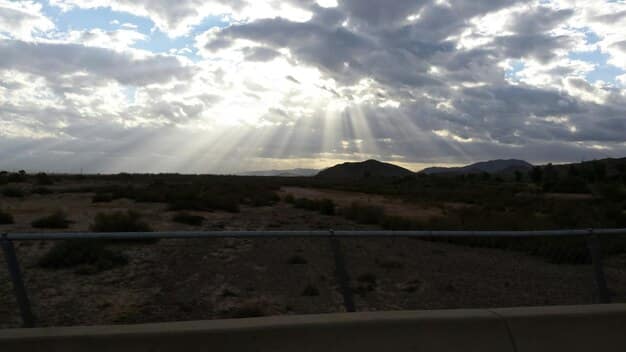A scenic overlook shot from a low angle, emphasizing the vastness of a desert landscape with a winding road leading to the horizon. The composition uses the rule of thirds, placing a blurred vintage campervan in the foreground to add depth and storytelling. The sky is partially cloudy, adding texture and drama to the scene, enhancing the overall sense of adventure and exploration.