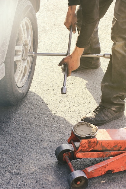 A roadside assistance worker changing a flat tire on a vehicle. The worker is wearing a reflective vest and using a lug wrench to remove the lug nuts from the tire. The car is parked on the side of a highway with other vehicles passing by.